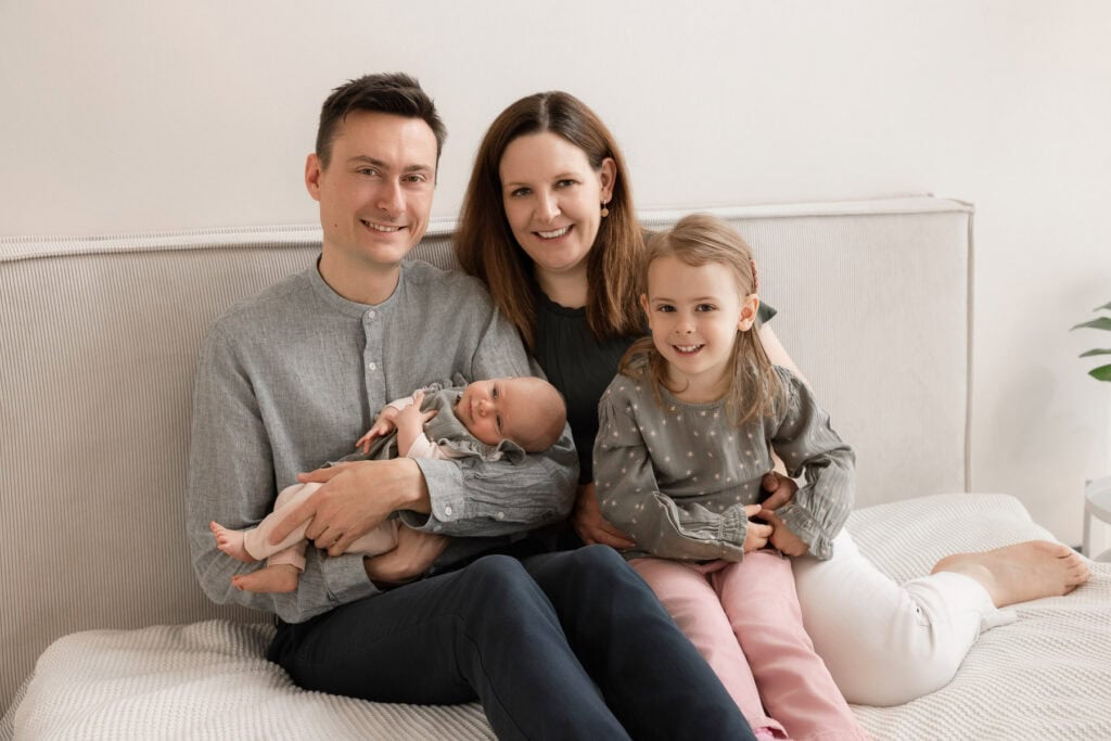 Familienfoto mit Eltern, Tochter und Neugeborenem auf dem Bett in gemütlicher Atmosphäre.